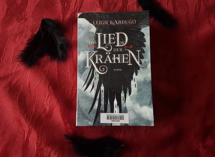 Das Buch "Das Lied der Krähen" liegt auf rotem Untergrund. Außenherum liegen schwarze Federn.