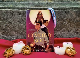 Das Buch "Cemetery Boys" steht vor einer dunklen Steinwand auf einem roten Untergrund. Davor stehen Kerzen und goldene Blüten. Am Buch hängt ein trans-Pride-Armband.