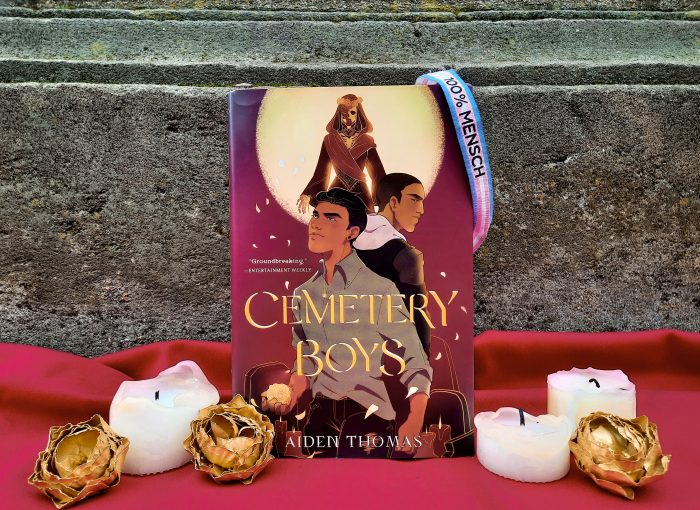 Das Buch "Cemetery Boys" steht vor einer dunklen Steinwand auf einem roten Untergrund. Davor stehen Kerzen und goldene Blüten. Am Buch hängt ein trans-Pride-Armband.
