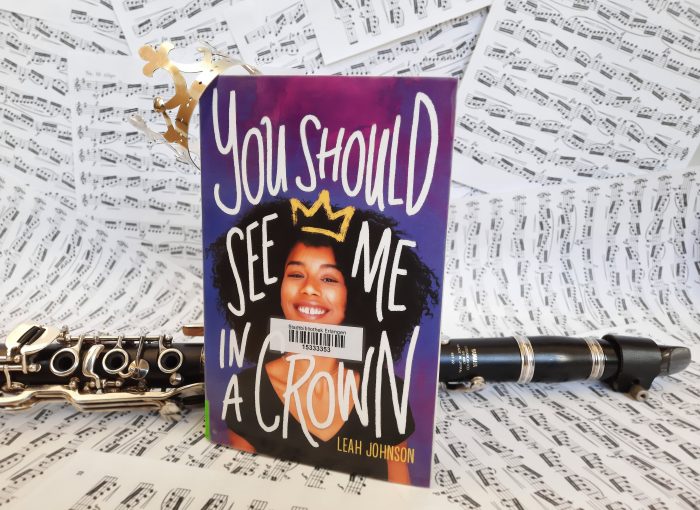 Das Buch "You should see me in a crown" steht an eine Klarinette gelehnt vor einem Hintergrund aus Notenblättern. Das Buch trägt ein Krönchen.