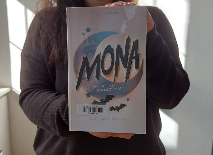 Ein Mensch hält das Buch "Mona: Und täglich grüßt der Erzdämon"