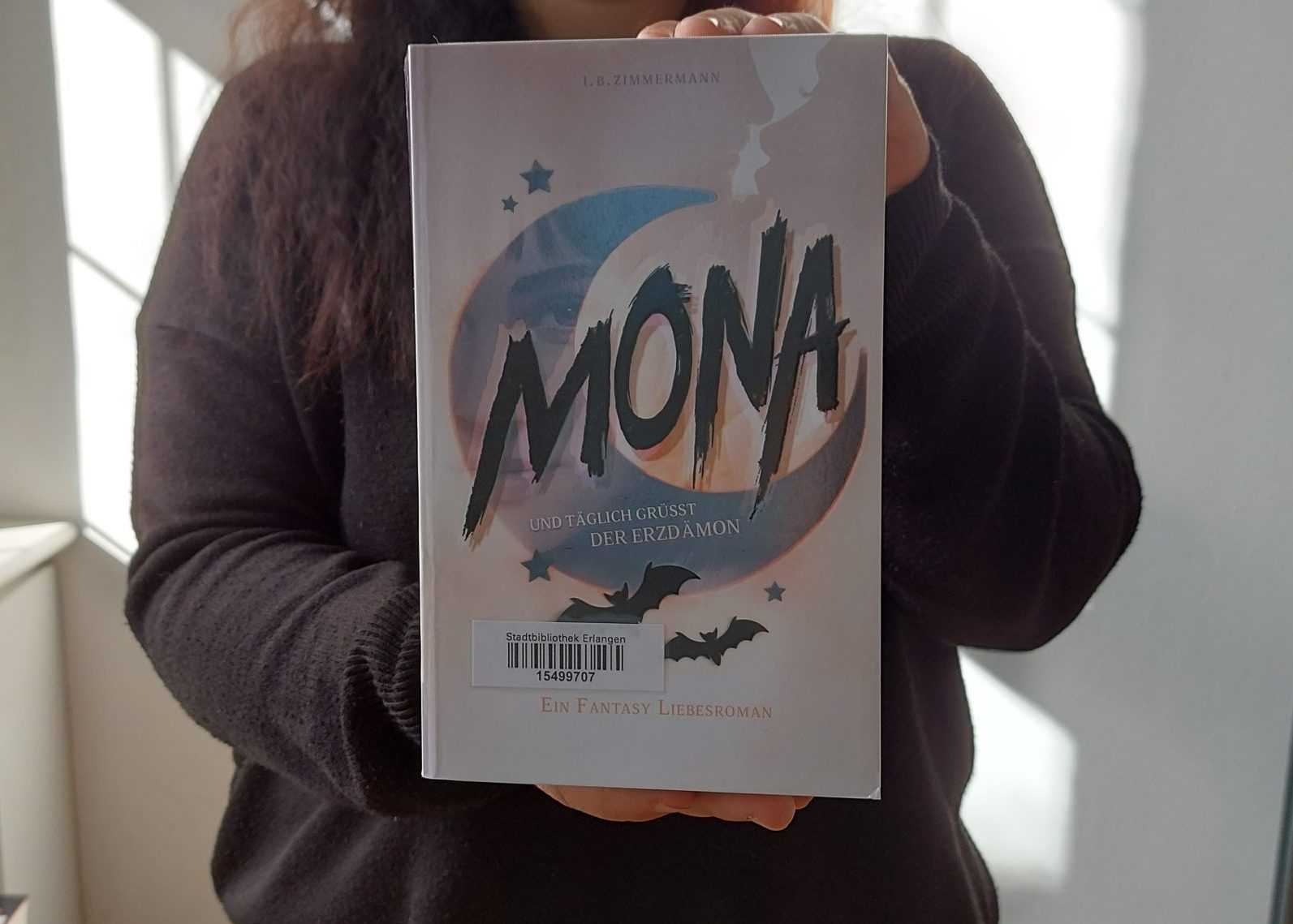 Ein Mensch hält das Buch "Mona: Und täglich grüßt der Erzdämon"