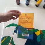 Das Buch "Das Café am Rande der Welt" von John Strelecky wird vor eine Leinwand mit Farbe gehalten. Diese wird mit einem Pinsel passend zum Buchcover gemalt.