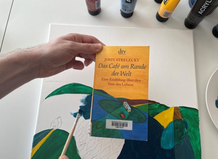 Das Buch "Das Café am Rande der Welt" von John Strelecky wird vor eine Leinwand mit Farbe gehalten. Diese wird mit einem Pinsel passend zum Buchcover gemalt.